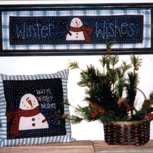 May include: A framed blue and white plaid fabric with the text "Winter Wishes" and a snowman with a red scarf. A pillow with a blue and white striped border and a snowman with a red scarf and the text "Warm Winter Wishes". A basket filled with pine branches.