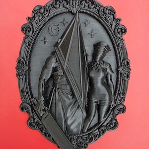 May include: A black oval wall plaque featuring a relief sculpture of a pyramid-headed figure holding a large blade next to a nurse figure. The background includes a crescent moon and star symbols, all enclosed within an ornate, decorative frame.