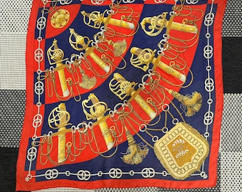 May include: A red and blue silk scarf with a gold and white pattern of swords and chains. The scarf has a gold border with a repeating pattern of circles and chains. The scarf has the text "Hermès Paris" in the center.