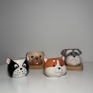 Puede incluir: Cuatro macetas de cerámica con forma de cara de perro. Las macetas son de color negro, blanco, marrón y naranja. Las macetas están sobre una superficie de madera.
