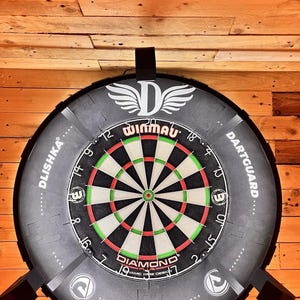 Op de afbeelding: Een Winmau Diamond Plus dartbord met een zwarte en grijze rand, gemonteerd op een houten muur. Het dartbord heeft een klassiek ontwerp met rode, groene en zwarte segmenten. De tekst "Winmau" en "Diamond" zijn zichtbaar.
