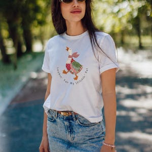 May include: A white short-sleeved t-shirt featuring a graphic of a goose with a patchwork-style pattern in red, green, and white. The text "Lookin' For My Stitchin' Pal" is printed in a circular arc around the goose illustration on the front.