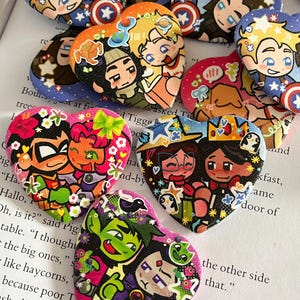 May include: A collection of heart-shaped pins featuring colourful cartoon illustrations of various fictional character pairs. Each pin displays distinct characters with vibrant details, including stars, flowers, and symbols, arranged on a printed book page background.