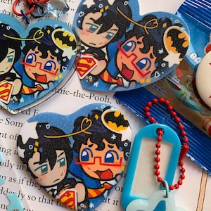 May include: Three heart-shaped acrylic charms featuring a cartoon illustration of two characters, one with black hair and one with curly hair wearing red glasses. The charms include a red lobster clasp, a blue plastic tag, and a red ball chain.
