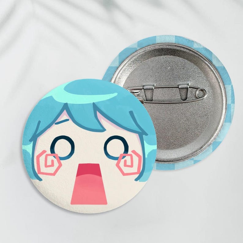 May include: Two round pinback buttons. One button features a cartoon face with blue hair, large black eyes, and a surprised expression. The other button shows the pinback mechanism. The background of the pin is light blue and white.