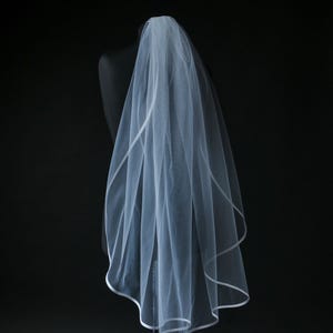 Può includere: Un velo da sposa bianco con bordo in raso, drappeggiato su un manichino nero. Il velo è realizzato in un tessuto trasparente e leggero, e il bordo segue il bordo del velo. Lo sfondo è nero.