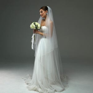 May include: A bride wearing a white strapless wedding dress with a long veil and holding a bouquet of white flowers.