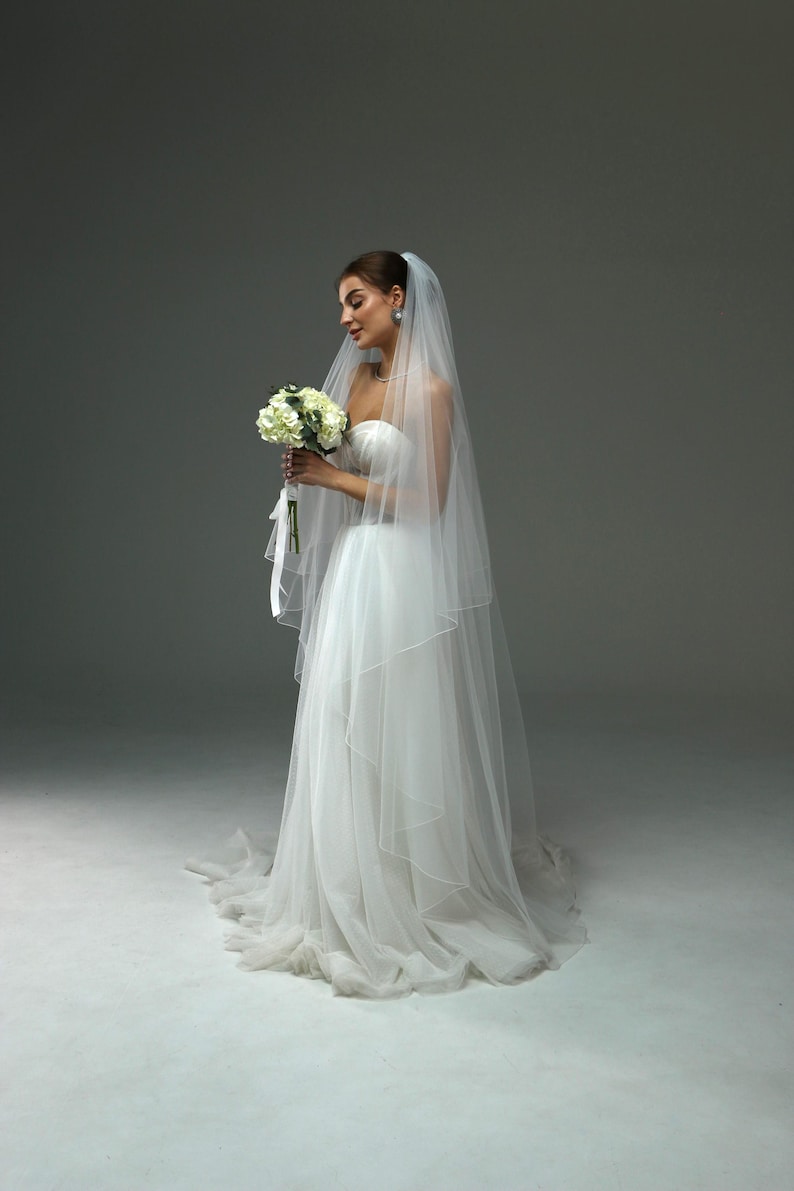 May include: A bride wearing a white strapless wedding dress with a long veil and holding a bouquet of white flowers.