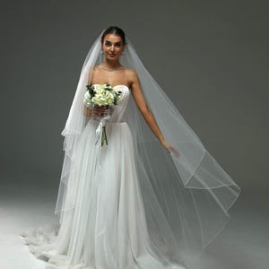 May include: A bride wearing a white strapless wedding dress with a long veil and holding a bouquet of white flowers.