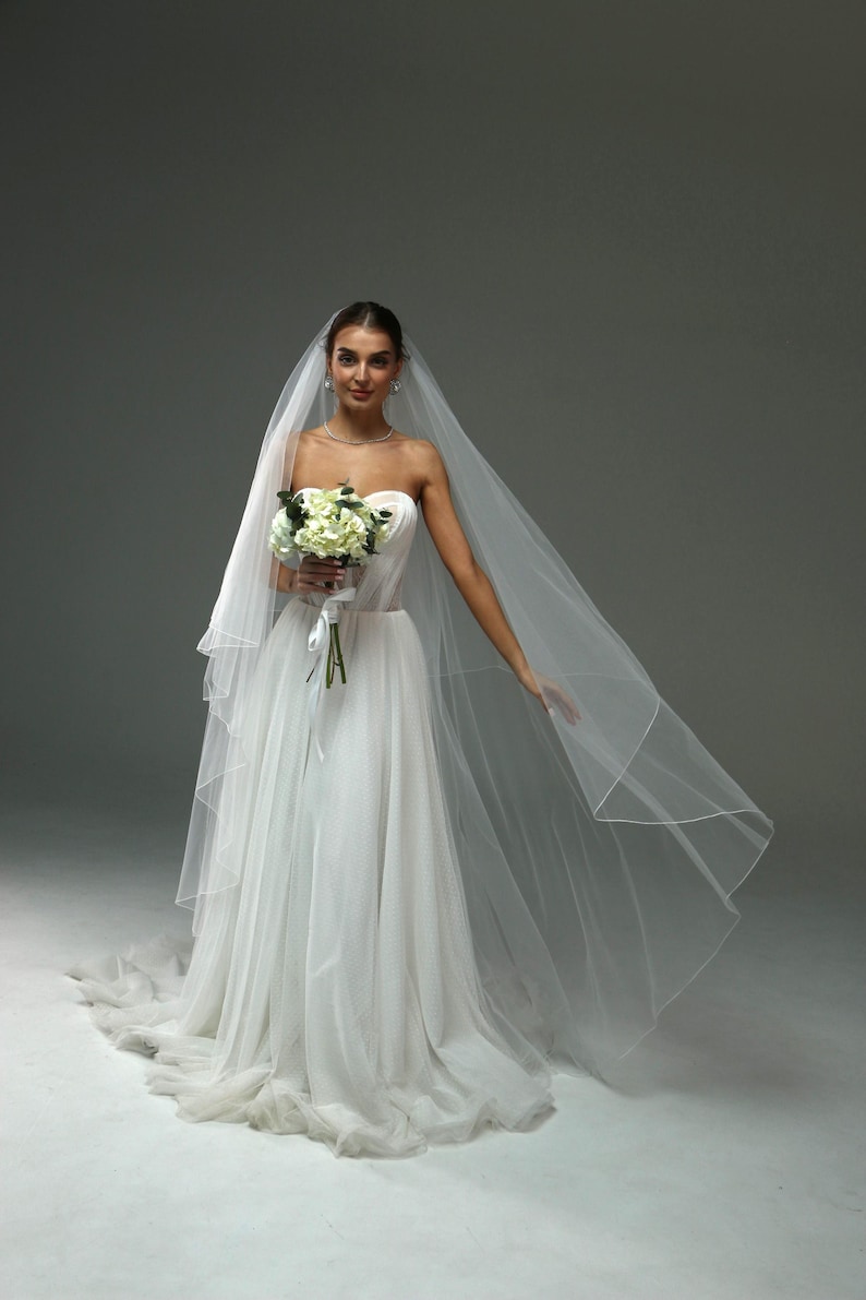 May include: A bride wearing a white strapless wedding dress with a long veil and holding a bouquet of white flowers.