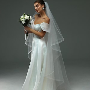 May include: A bride wearing a white off-the-shoulder wedding dress with a long veil and holding a bouquet of white flowers.