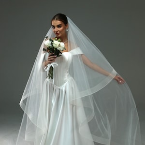 Pode incluir: Uma noiva vestindo um vestido de noiva branco com um véu longo e segurando um buquê de flores brancas.