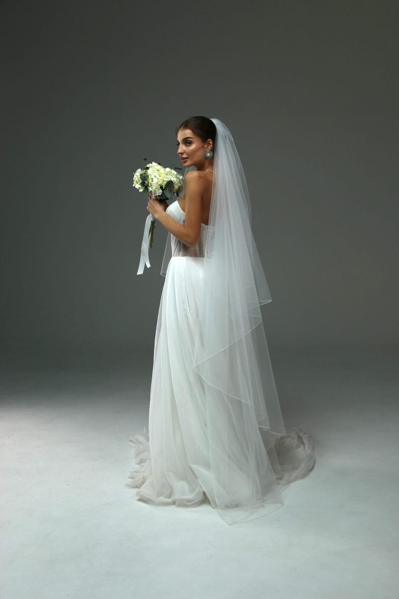 May include: A bride wearing a white wedding dress with a long veil and holding a bouquet of white flowers.