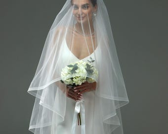 May include: A bride wearing a white wedding dress and a long white veil holds a bouquet of white flowers.