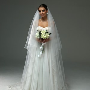 May include: A bride wearing a white wedding dress with a long veil and holding a bouquet of white flowers.