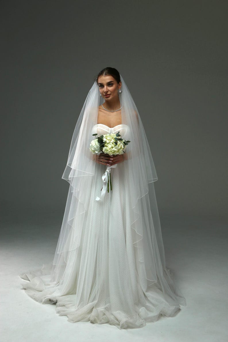 May include: A bride wearing a white wedding dress with a long veil and holding a bouquet of white flowers.