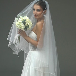 May include: A bride wearing a white wedding dress with a long, white veil. She is holding a bouquet of white flowers.