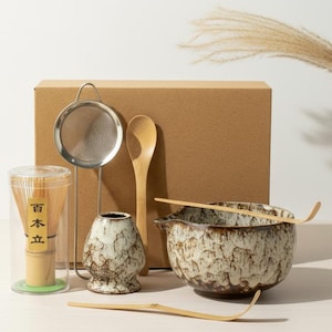 May include: A matcha tea set includes a ceramic bowl, bamboo whisk, scoop, and spoon, along with a stainless steel strainer and a small ceramic container. The set is presented with a cardboard box in the background. The whisk is in a clear container with Japanese text.