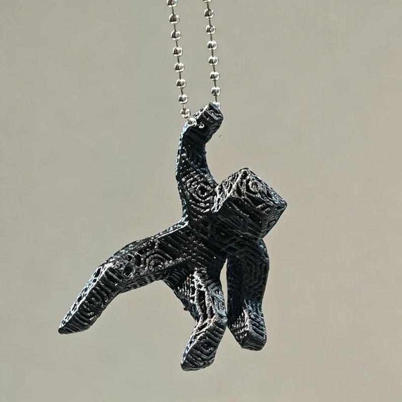 May include: A black pendant necklace featuring a stylized human figure. The pendant is intricately detailed with a textured surface and hangs from a silver ball chain. The figure is in a dynamic pose, suggesting movement.