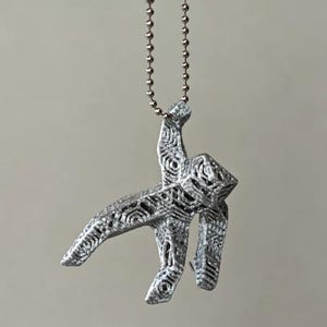 May include: A silver-colored pendant in the shape of a stylized figure, suspended from a ball chain. The pendant has an open, geometric design with intricate patterns. The background is a solid, neutral color.