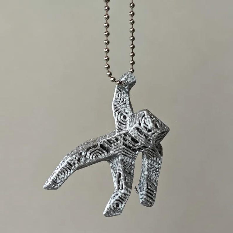 May include: A silver-colored pendant in the shape of a stylized figure, suspended from a ball chain. The pendant has an open, geometric design with intricate patterns. The background is a solid, neutral color.