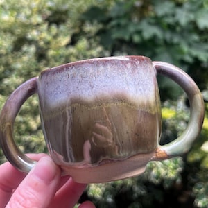 May include: A ceramic mug featuring two curved handles on opposite sides. The exterior has a reactive glaze finish with a gradient transition from a light, creamy white at the top rim to a deep, earthy brown and muted green tone at the base.