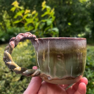 May include: A ceramic mug featuring a braided handle and a reactive glaze finish. The mug displays a gradient of earthy green and brown tones, with a smooth, glossy texture that drips down the sides towards the unglazed, natural clay base.