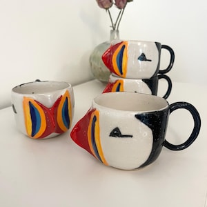 May include: A collection of ceramic mugs shaped like puffins, featuring white bodies with black accents, and prominent, colourful beaks painted in red, yellow, and blue. The mugs have black handles and are arranged in a group on a white surface.