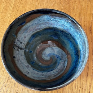 May include: A round ceramic bowl featuring a glossy glaze with a swirling pattern of deep navy blue, bright sky blue, and earthy brown tones. The interior surface displays a smooth, reflective finish with a distinct spiral design radiating from the centre.