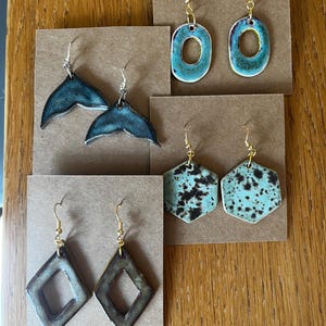 May include: Four pairs of ceramic earrings on brown display cards. One pair is teal oval hoops, one is dark teal whale tails, one is light blue with black speckles in a hexagon shape, and one is dark brown diamond-shaped open frames with gold hooks.