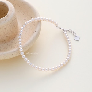 May include: A delicate pearl bracelet with a silver clasp and a small heart charm. The bracelet is composed of numerous small, round, white pearls. The bracelet is displayed on a cream-colored surface, next to a ceramic cup and plate.