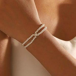 Può includere: Un braccialetto di perle a due fili con un piccolo elemento centrale quadrato a forma di diamante. Il braccialetto è indossato al polso di una persona, con i fili di perle che si incurvano attorno al braccio. Le perle sono bianche e l'elemento centrale è trasparente.