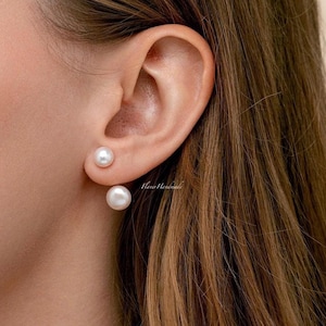 May include: Close-up of an ear adorned with two white pearl earrings. The earrings are spherical and have a glossy finish. The ear is set against a backdrop of brown hair.