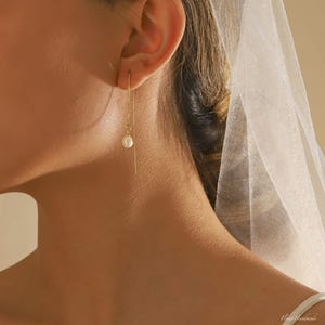 Può includere: Primo piano di un orecchio con un delicato orecchino a catena dorata con una piccola perla bianca.