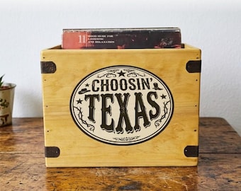 Może przedstawiać: Drewniana skrzynia do przechowywania z napisem "CHOOSIN' TEXAS" w czarno-białym owalnym wzorze. Skrzynia ma metalowe wzmocnienia narożne i mieści album. Skrzynia stoi na drewnianej powierzchni.