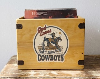 Może przedstawiać: Drewniane pudełko do przechowywania z naklejką o tematyce kowbojskiej. Na naklejce widnieje napis "God Loves Cowboys" i przedstawia kowboja na koniu. Pudełko zawiera album płytowy.