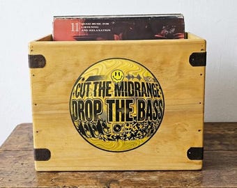 Może przedstawiać: Drewniana skrzynia do przechowywania płyt z żółto-czarną grafiką z napisem "CUT THE MIDRANGE DROP THE BASS". Skrzynia ma metalowe wzmocnienia narożne i mieści album z tekstem "Good Music for Listening and Relaxation".