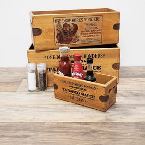 May include: Wooden crates with the text "ONE DROP WORKS WONDERS" and "TABASCO SAUCE" are stacked. The crates contain bottles of sauce, salt, and pepper. The image has a vintage aesthetic.