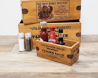 Może przedstawiać: Drewniane skrzynie z napisem "ONE DROP WORKS WONDERS" i "TABASCO SAUCE" są ułożone w stos. Skrzynie zawierają butelki z sosem, solą i pieprzem. Obraz ma estetykę vintage.