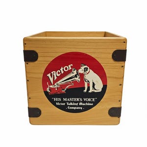 May include: Light brown wooden box with a vintage "Victor Talking Machine Company" logo. The logo features a dog listening to a gramophone. Dark metal corner reinforcements.