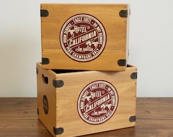 Może przedstawiać: Dwie drewniane skrzynie do przechowywania ułożone jedna na drugiej. Każda skrzynia ma okrągły emblemat z napisem "Hotel California Los Angeles Pink Champagne Only". Skrzynie są jasnobrązowe z czarnymi wzmocnieniami narożników.