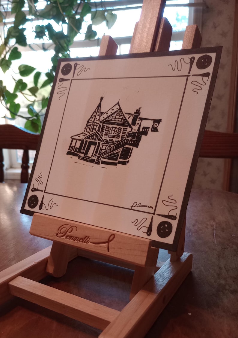 May include: A black and white linocut print of a house with a decorative border. The print is on a white background and is mounted on a wooden easel. The easel has the name "Pennell" engraved on it.