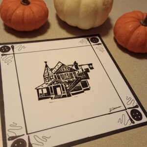 May include: Black and white linocut print of a house with a decorative border of buttons and thread. The print is signed "J.C. Cowan".