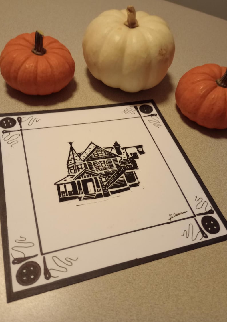 May include: Black and white linocut print of a house with a decorative border of buttons and thread. The print is signed "J.C. Cowan".