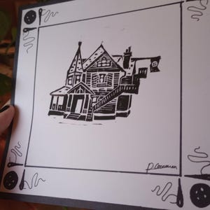 May include: Black and white linocut print of a house with a porch and a flag on the roof. The print is framed with a black border and has a signature in the bottom right corner.