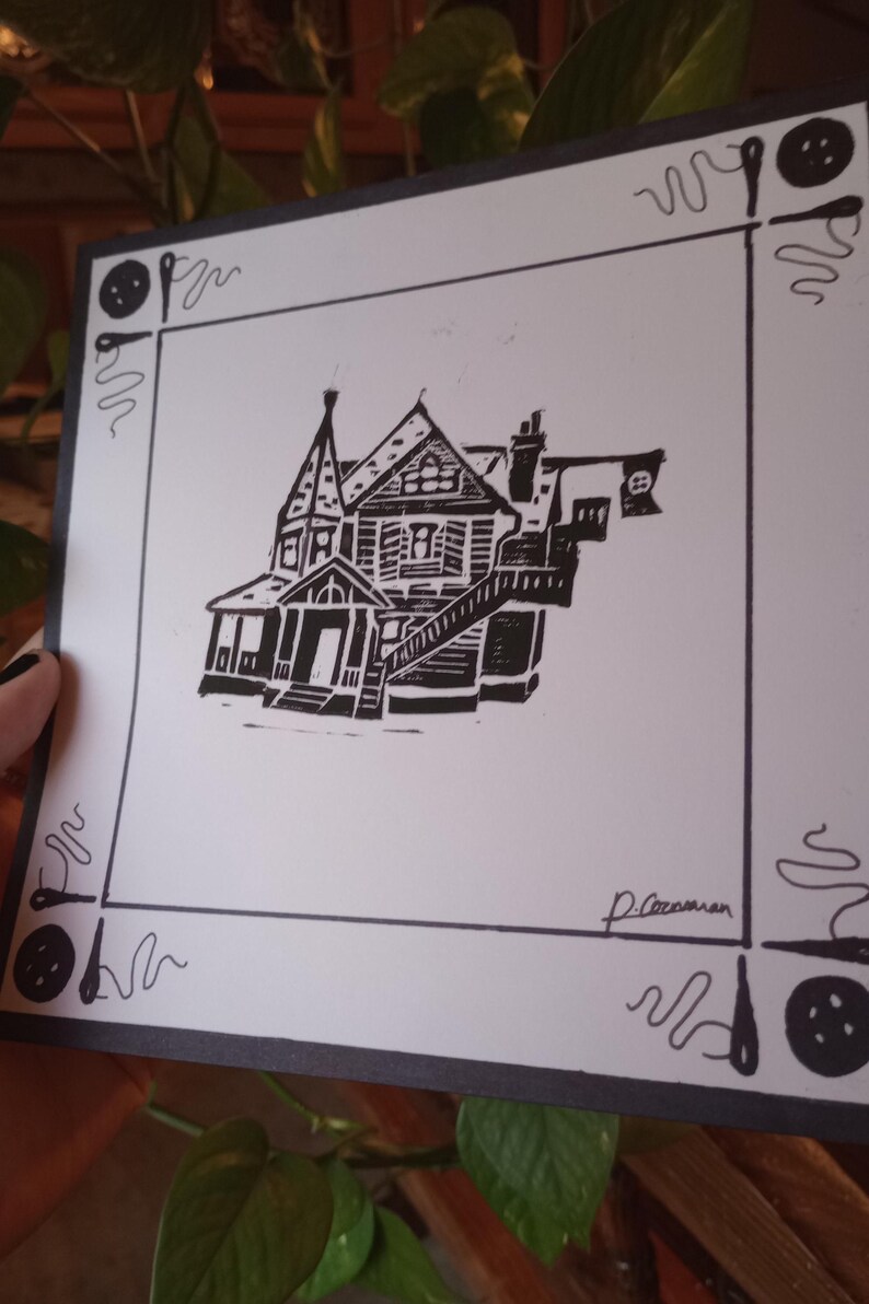 May include: Black and white linocut print of a house with a porch and a flag on the roof. The print is framed with a black border and has a signature in the bottom right corner.