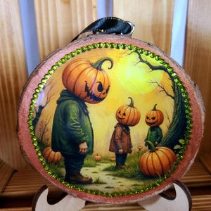 May include: A round wooden Halloween ornament with a painted scene of pumpkin-headed figures in a spooky landscape. The ornament has a green gem border and a black ribbon. The artwork features a yellow sky and orange pumpkins.