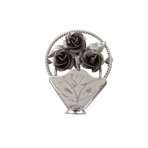 Peut inclure: Broche argentée représentant trois roses dans un cadre circulaire, au-dessus d'une base en forme de panier. La broche présente un design détaillé et texturé, avec des motifs complexes sur les roses et le panier. Le fond est blanc.