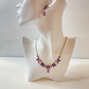 Peut inclure: Un ensemble collier et boucles d'oreilles argenté avec des motifs floraux violets et bleus. Le collier présente des grappes en forme de fleurs et un pendentif en forme de goutte. Les boucles d'oreilles ont un design floral assorti.