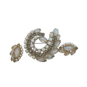 Peut inclure: Un ensemble de bijoux vintage comprenant un broche et des boucles d'oreilles assorties à clip. La broche est en forme de croissant et les boucles d'oreilles sont ovales. L'ensemble est orné de pierres blanches et vert olive.